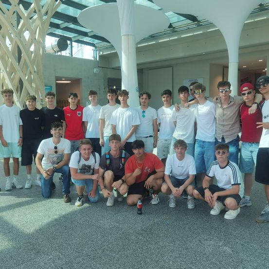 Eine Gruppe von Teenagern posiert für ein Foto in einem Innenbereich, einige mit Brillen und Mützen, einer hält eine Flasche. Sie lächeln und stehen eng beieinander.