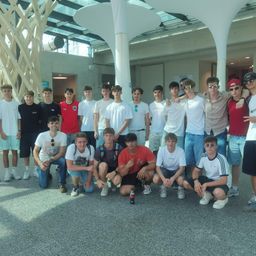Eine Gruppe von Teenagern posiert für ein Foto in einem Innenbereich, einige mit Brillen und Mützen, einer hält eine Flasche. Sie lächeln und stehen eng beieinander.