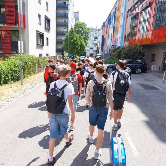 Eine Gruppe junger Leute geht mit ihren Rucksäcken und Gepäck die Straße entlang. Die Straße ist von Gebäuden und Grünflächen gesäumt.