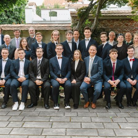 Eine Gruppe formell gekleideter Personen, darunter Männer und Frauen, sitzen auf einer Bank und posieren für ein Foto. Der Hintergrund zeigt eine Außenumgebung mit einer Backsteinmauer und Grünpflanzen.