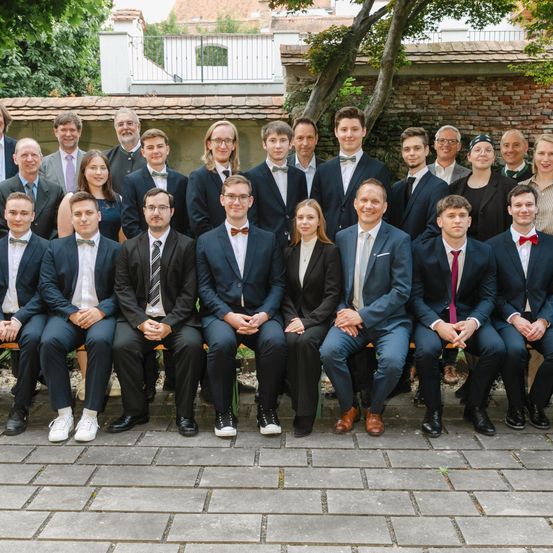 Eine Gruppe formell gekleideter Personen, darunter Männer und Frauen, sitzen auf einer Bank und posieren für ein Foto. Der Hintergrund zeigt eine Außenumgebung mit einer Backsteinmauer und Grünpflanzen.