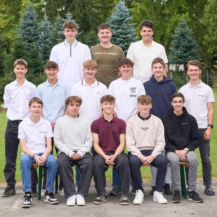 Eine Gruppe junger Männer posiert für ein Klassenfoto in einem Park, in verschiedenen Freizeit- und Sportkleidung. Sie sitzen auf Bänken mit Bäumen und Gras im Hintergrund.