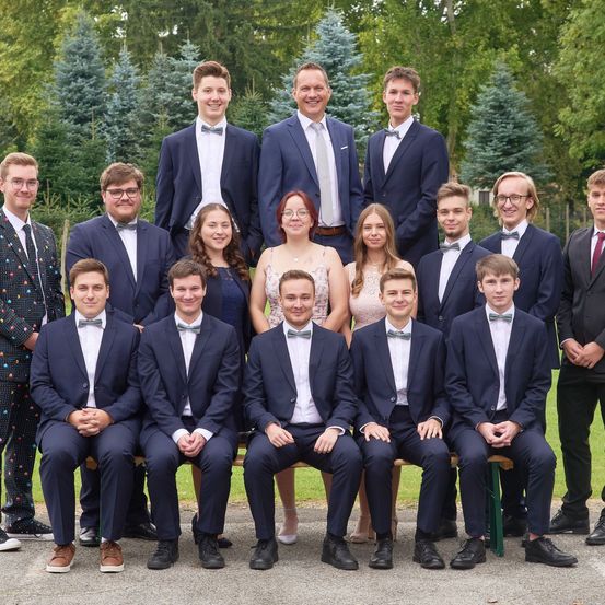 Eine Gruppe von Menschen, darunter Schüler und Lehrer, posiert für ein Foto in einem Park. Sie sind in formellen Anzügen und Krawatten gekleidet, einige tragen Brillen. Der Hintergrund besteht aus Bäumen.