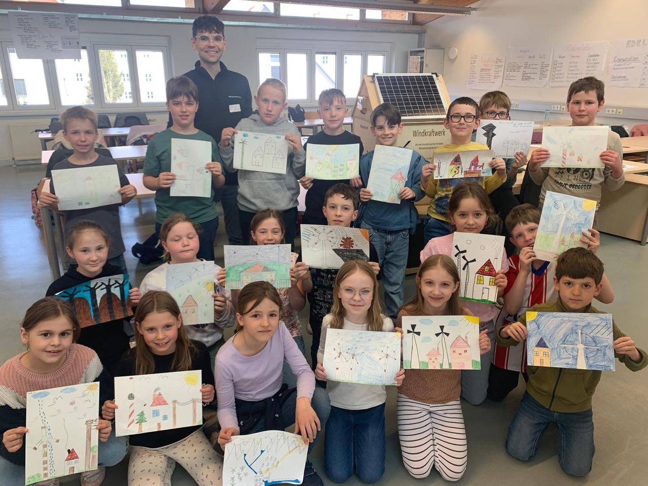 Eine Gruppe von Kindern in einem Klassenzimmer posiert mit ihren Zeichnungen. Sie halten Papiere mit Zeichnungen von Häusern, Bäumen und Solarpanelen. Die Kinder lächeln und schauen in die Kamera.