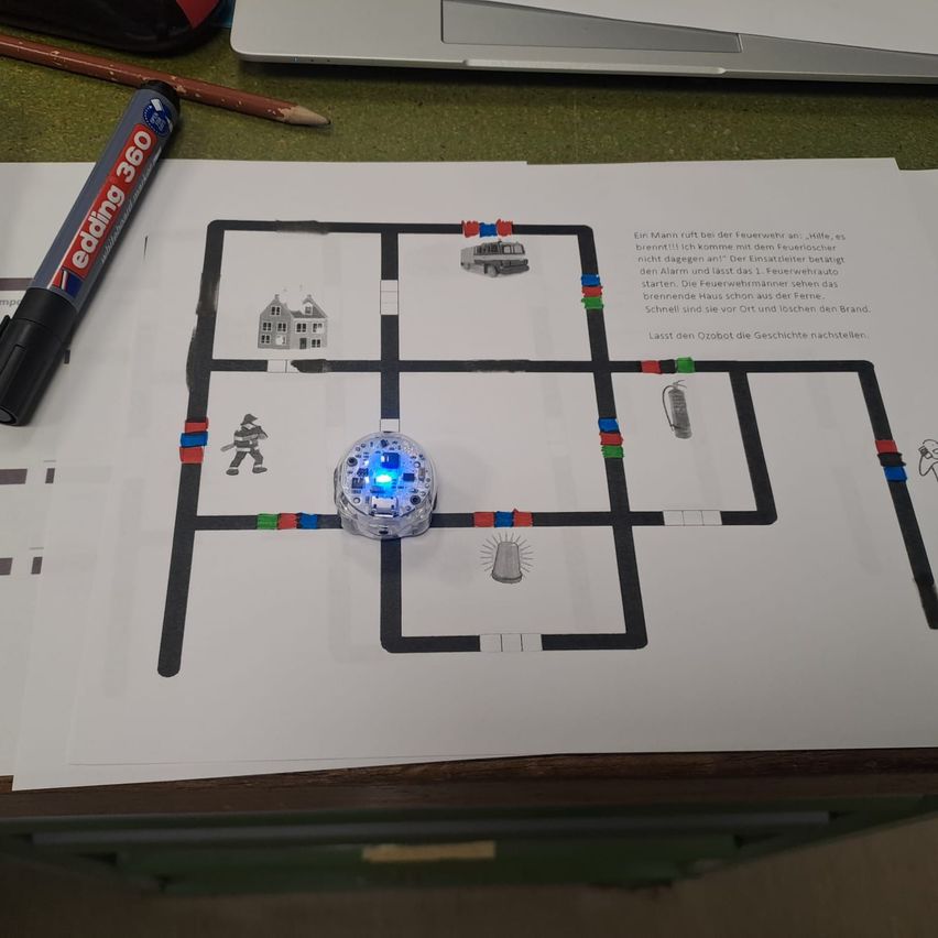 Eine Papierzeichnung eines Labyrinths mit einem Roboter darauf, mit einem Feuerlöscher und einem Haus, zusammen mit einem Marker und einem Bleistift auf einem Schreibtisch.