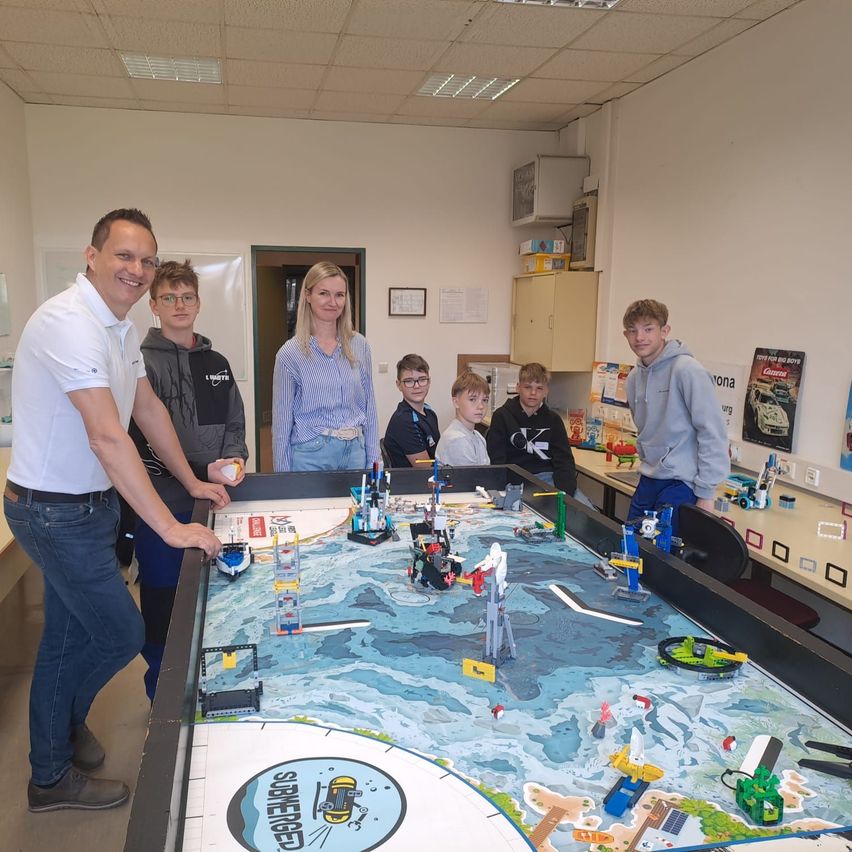 Drei Erwachsene und vier Kinder stehen um einen großen Tisch in einem Raum. Der Tisch hat eine blaue Karte mit mehreren Lego-Stücken. Die Kinder schauen sich die Lego-Stücke an.