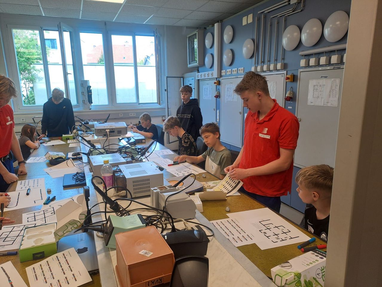 In einem Klassenzimmer arbeiten eine Gruppe von Schülern unter Anleitung eines Lehrers an einem Elektronikprojekt. Der Tisch ist mit elektronischen Komponenten, Kabeln und Papieren übersät.