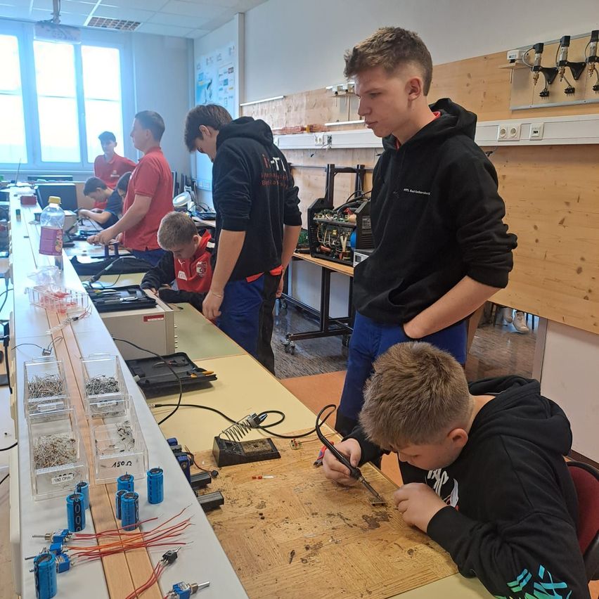 Eine Gruppe von Jungen arbeitet in einem Labor, wobei ein Junge ein Löteisen auf einer Holzplatte benutzt, ein anderer steht und andere an langen Tischen mit verschiedenen elektronischen Komponenten und Werkzeugen sitzen.