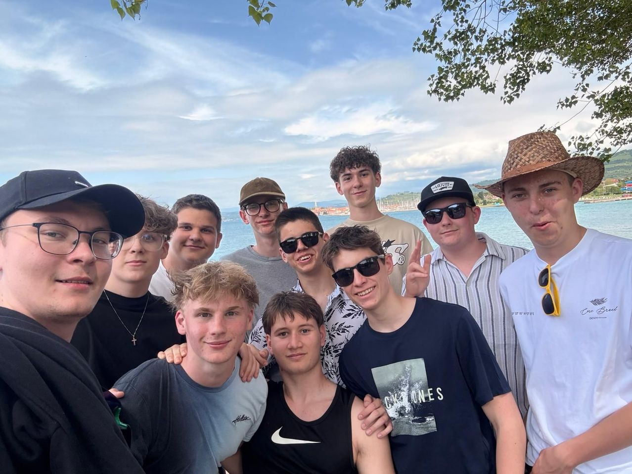 Eine Gruppe junger Männer posiert für ein Foto in der Nähe eines Sees. Einige tragen Sonnenbrillen und Hüte. Der Hintergrund zeigt einen bewölkten Himmel und eine entfernte Stadt.