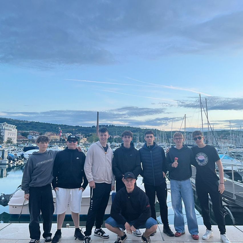 Eine Gruppe junger Männer steht bei Sonnenuntergang auf einem Steg, mehrere Boote sind dahinter verankert und der Himmel ist bewölkt.