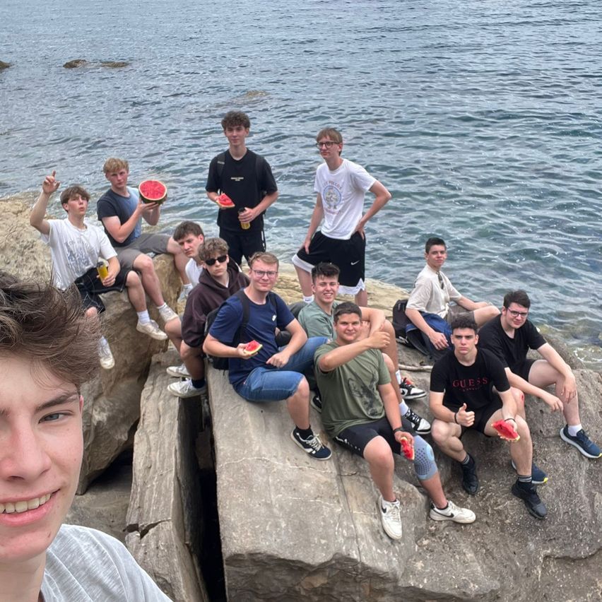 Eine Gruppe von Teenagern posiert für ein Foto auf Felsen am Meer und hält Wassermelonenstücke in der Hand. Das Meer ist ruhig und der Himmel ist klar.