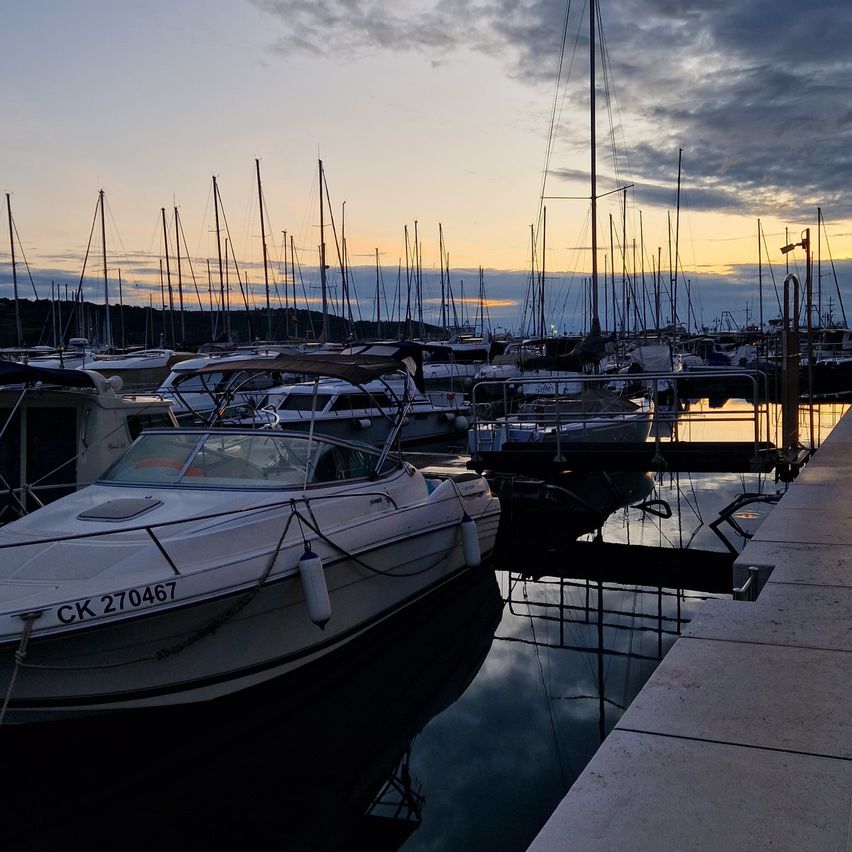 Eine Marina bei Sonnenuntergang mit zahlreichen verankerten Segelbooten. Ein weißes Boot mit der Aufschrift CK 270467 ist im Vordergrund und spiegelt sich im Wasser.
