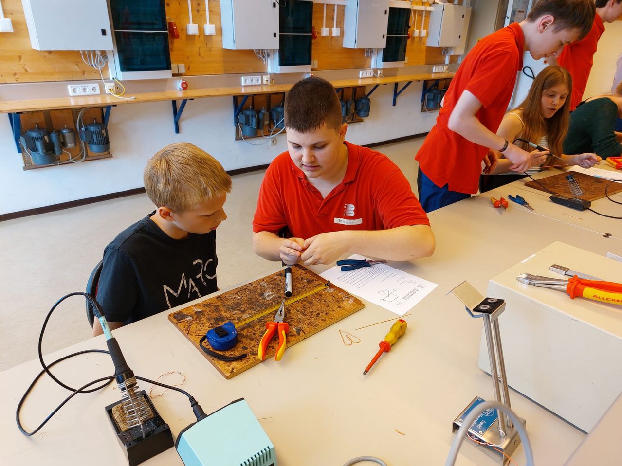 Zwei Jungen in einer Bildungseinrichtung, einer in einem roten Shirt arbeitet an einem Lötprojekt, während ein anderer zusieht. Verschiedene Werkzeuge und Materialien sind auf einem Tisch verteilt.