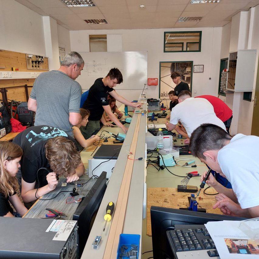 Eine Gruppe von Menschen arbeitet in einem Raum mit weißen Wänden und einer Tafel an elektronischen Geräten. Einige löten, während andere Werkzeuge auf dem langen Tisch verwenden.