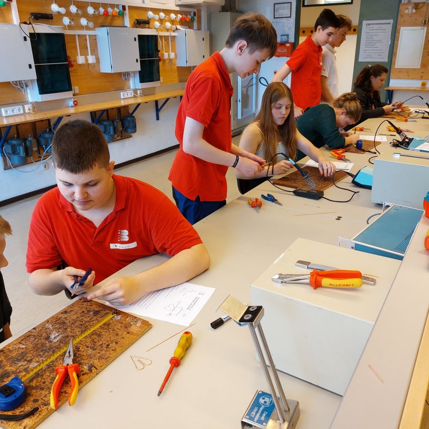 Eine Gruppe von Schülern in roten Hemden arbeitet in einem Labor an einem Projekt. Sie verwenden Werkzeuge und Kabel.