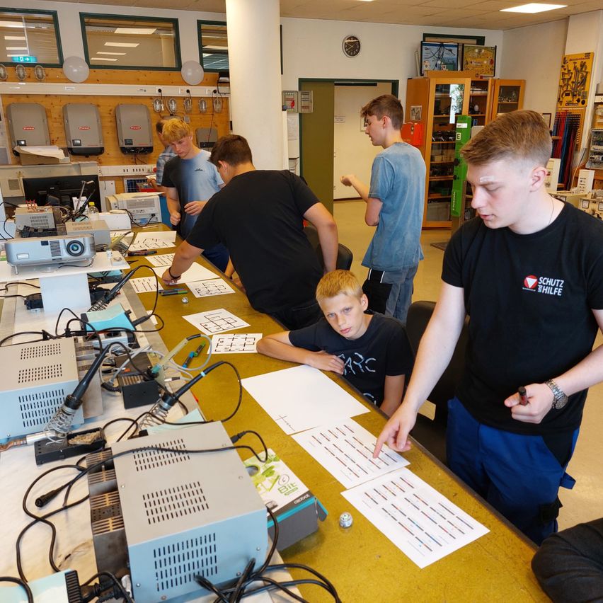 Mehrere junge Männer arbeiten in einem Labor und stehen um einen Tisch voller elektronischer Geräte. Ein Mann zeigt auf ein Diagramm auf dem Tisch.