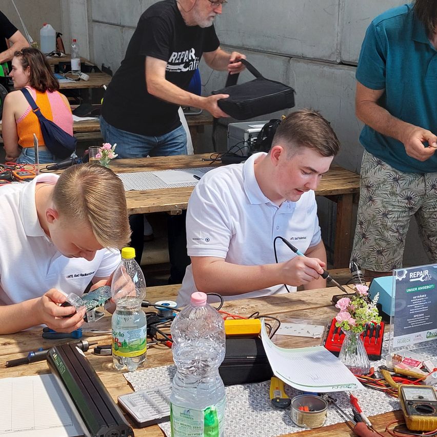 Zwei junge Männer arbeiten an einem elektronischen Projekt am Tisch. Einer hält ein Lötkolben, während der andere an einer Leiterplatte arbeitet. Ein Mann mit Brille und Tasche steht in der Nähe.
