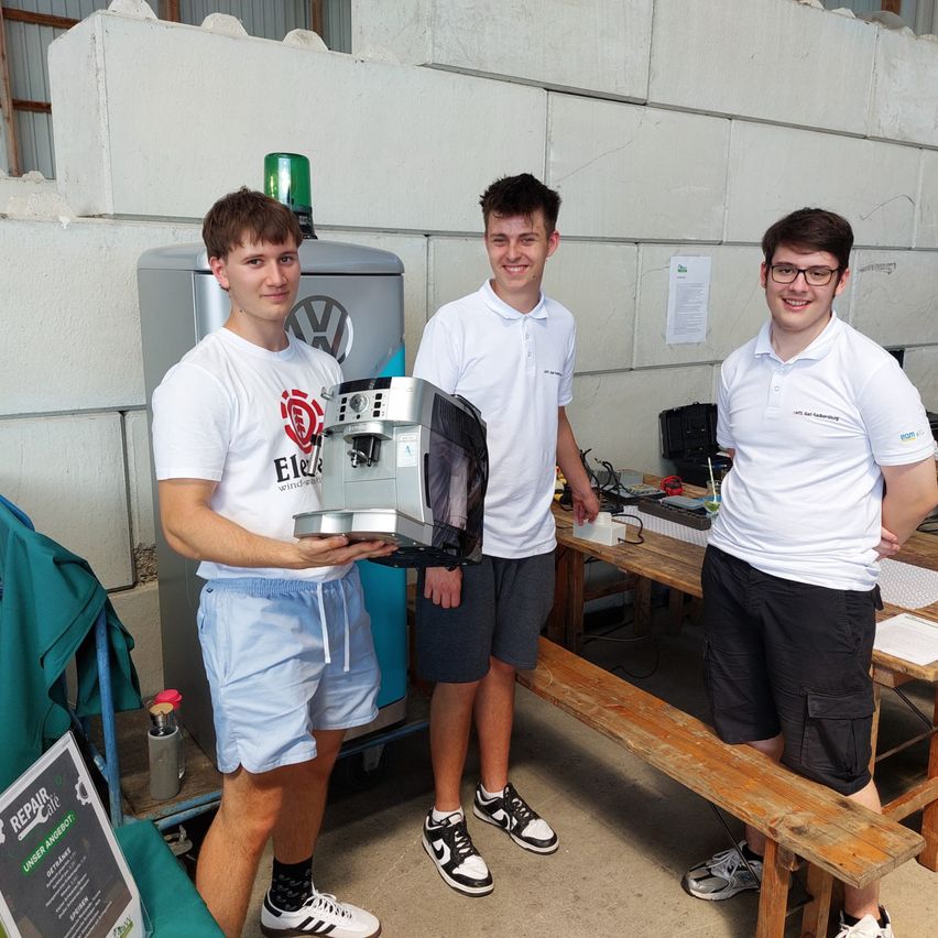 Drei junge Männer stehen draußen neben einer großen Maschine. Zwei tragen eine Brille, einer trägt Turnschuhe. Hinter ihnen eine weiße Wand mit einem Papier und einem grünen Licht. Holztische mit Gegenständen und eine Holzbank.