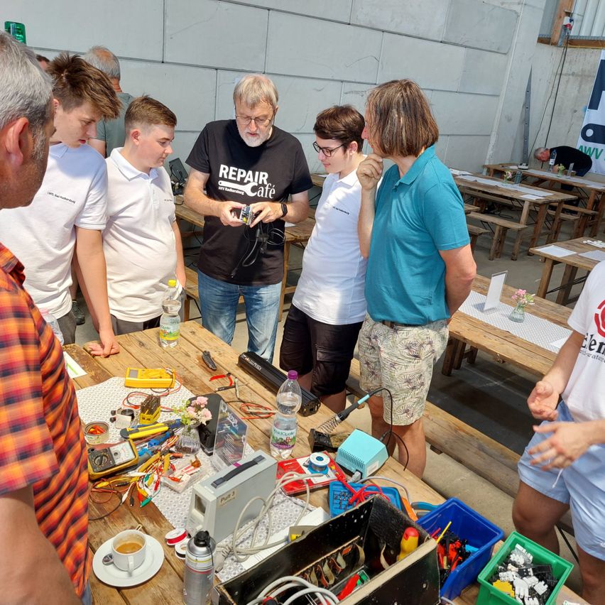 Eine Gruppe von Menschen ist um einen Tisch mit verschiedenen elektronischen Geräten versammelt, möglicherweise in einer Werkstatt. Ein Mann in einem schwarzen T-Shirt erklärt etwas an die Gruppe. Der Tisch ist mit Drähten, einem Multimeter und anderen Werkzeugen gefüllt. Die Umgebung scheint eine Werkstatt oder ein Reparaturcafé zu sein.