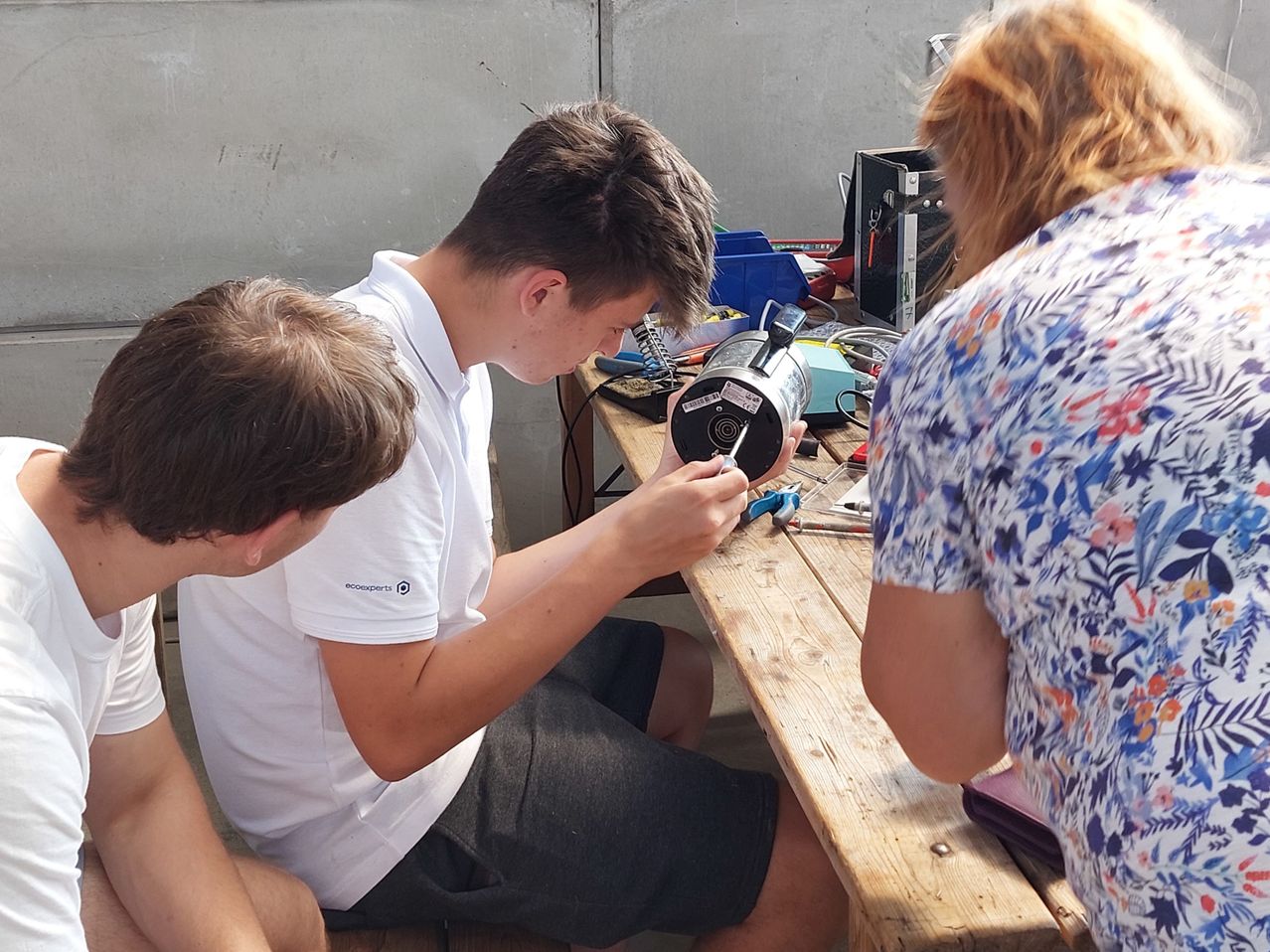 Drei Personen arbeiten an einem elektronischen Gerät an einem Holztisch. Ein Mann hält ein Werkzeug, während zwei andere beobachten.