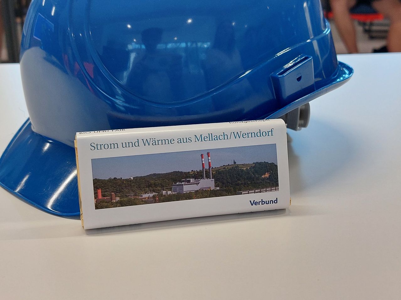 Eine kleine Schachtel steht vor einem blauen Helm. Die Schachtel trägt die Aufschrift 'Strom und Wärme aus Mellach/Wendorf' und zeigt ein Bild einer Fabrik.