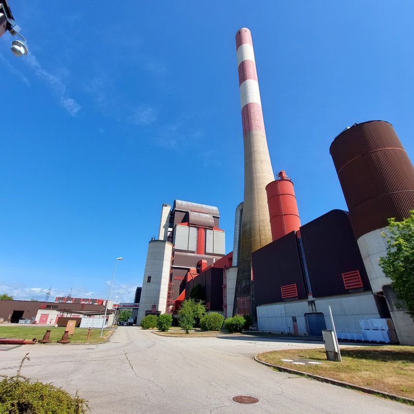 Ein Industriebetrieb mit hohen Schornsteinen und großen Tanks unter einem blauen Himmel. Eine breite Straße führt zum Werk.