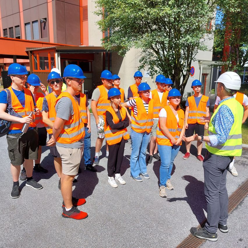 Eine Gruppe von Menschen in orangenen Westen und Helmen steht in einer Reihe draußen und diskutiert möglicherweise ein Thema, während ein Mann in einem weißen Helm zuhört.