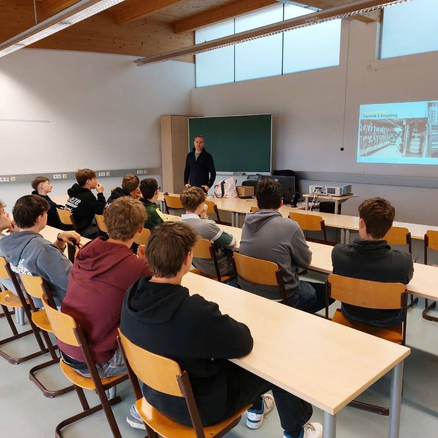 Ein Klassenzimmer mit Schülern an Tischen sitzend, ein Mann steht vor einer Projektionsleinwand und hält eine Vorlesung, dahinter eine Tafel.