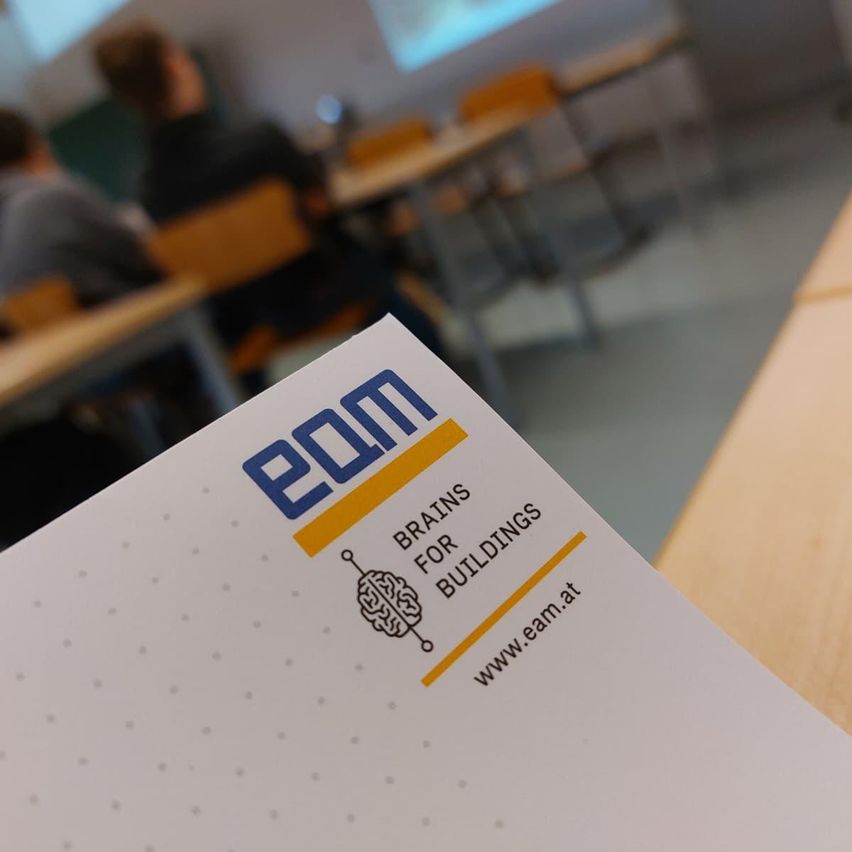 Ein Nahaufnahme einer weißen Visitenkarte mit 'EOM' und 'Brains for Buildings' sowie einer Website-Adresse. Der unscharfe Hintergrund zeigt einen Klassenraum mit sitzenden Personen.