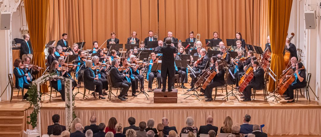 Ein Symphonieorchester spielt auf der Bühne mit einem Dirigenten, Geigern, Cellisten und Blechbläsern. Das Publikum sitzt in der ersten Reihe und schaut zu.
