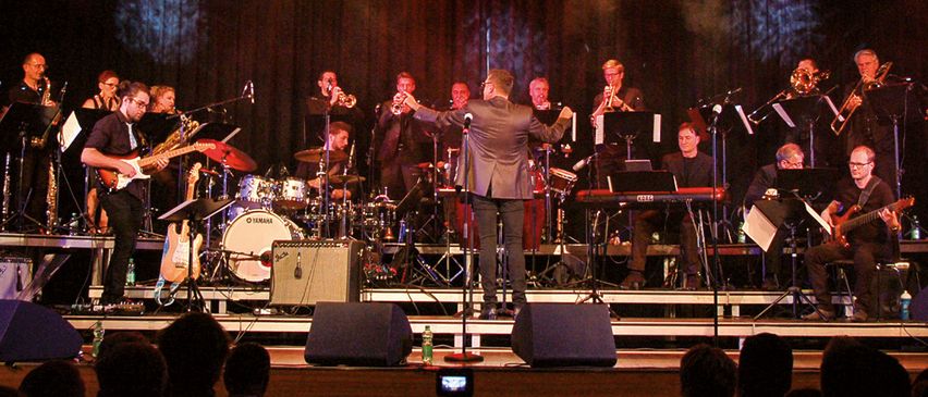 Eine Band spielt auf einer Bühne unter roten Lichtern mit Instrumenten wie Trompeten, Saxophonen und Schlagzeug. Das Publikum sitzt vor der Bühne.