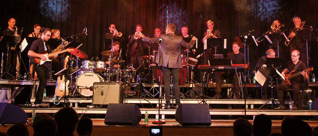 Eine Band spielt auf einer Bühne unter roten Lichtern mit Instrumenten wie Trompeten, Saxophonen und Schlagzeug. Das Publikum sitzt vor der Bühne.