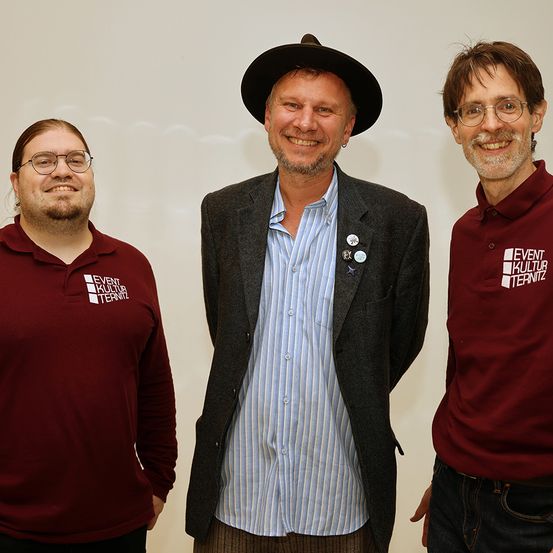 Drei Männer stehen zusammen und lächeln. Der Mann auf der linken Seite trägt ein Weinrot-Shirt mit weißem Text, der Mann in der Mitte ein blaues Hemd und einen grauen Mantel, der rechte Mann ein Weinrot-Shirt mit weißem Text.