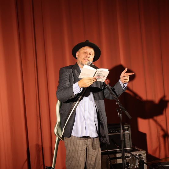 Ein Mann in einem Hut liest ein Buch in ein Mikrofon auf einer Bühne mit rotem Vorhang im Hintergrund.