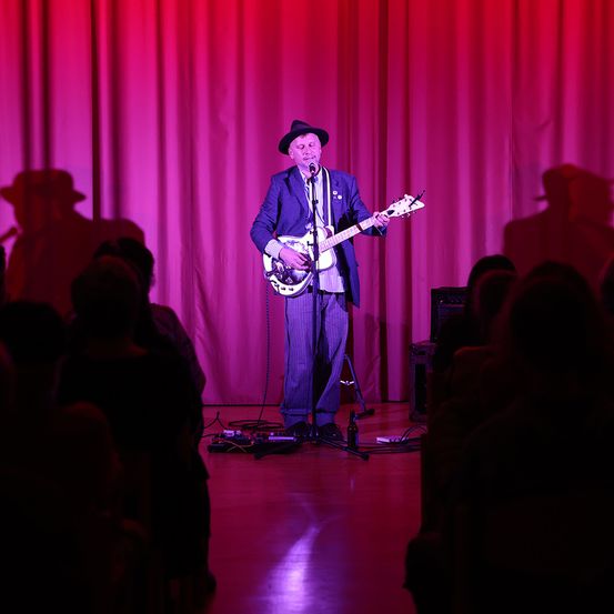 Ein Mann in Anzug und Hut spielt Gitarre auf der Bühne, mit rotem Vorhang im Hintergrund und Publikum davor.