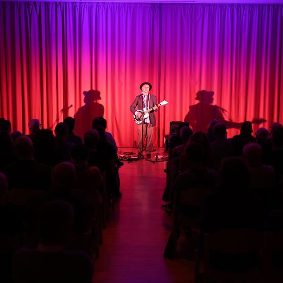 Ein Mann spielt auf der Bühne mit einer Gitarre. Die Bühne ist mit roten und lila Lichtern beleuchtet. Das Publikum sitzt vor ihm.