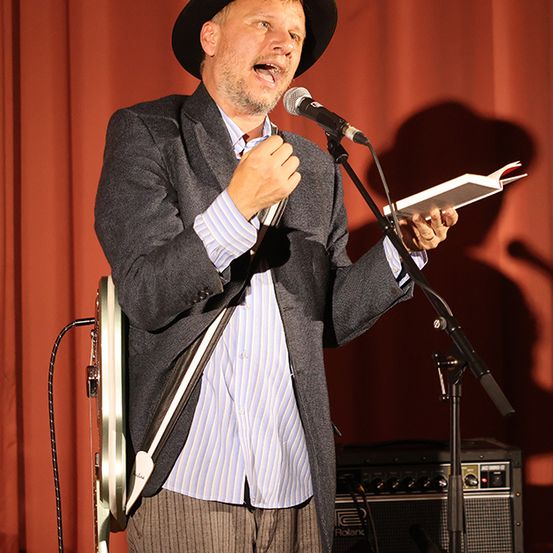 Ein Mann in einem schwarzen Hut und Anzug steht auf der Bühne, singt in ein Mikrofon und hält ein Buch. Hinter ihm ist ein roter Vorhang und ein Lautsprecher am Boden.