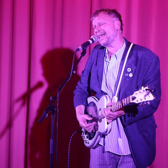Ein Mann tritt mit einer Gitarre auf der Bühne auf und singt in ein Mikrofon. Er trägt einen lilafarbenen Mantel und ein gestreiftes Hemd, beleuchtet von einem rosa Licht.