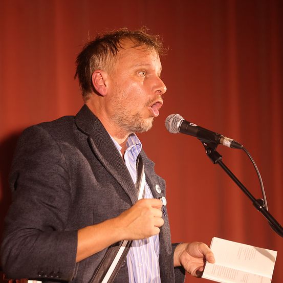 Ein Mann steht an einem Mikrofon und hält ein Buch. Er scheint eine Rede zu halten. Er trägt eine graue Jacke und ein gestreiftes Hemd. Hinter ihm ein roter Vorhang.