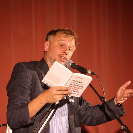 Ein Mann liest aus einem Buch mit dem Titel 'Wenn wir einmal nicht grausam sind, dann glauben wir gleich', während er vor einem Mikrofon steht. Er trägt einen dunklen Anzug und spricht leidenschaftlich.