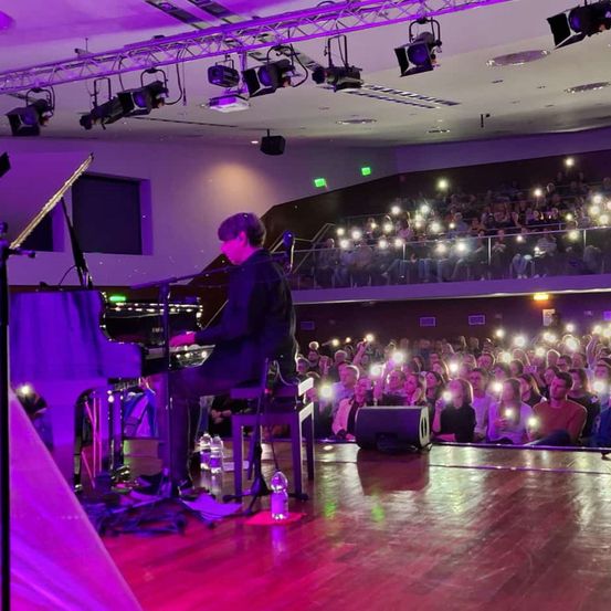 Ein Pianist sitzt auf einer Bühne unter lila Lichtern, mit einem großen Publikum im Raum, viele halten ihre Handys hoch.