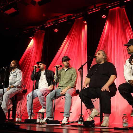 Fünf Männer sitzen auf Hockern auf einer Bühne. Sie treten mit Mikrofonen vor ihnen auf. Hinter ihnen sind rote Vorhänge und Bühnenbeleuchtung.