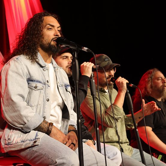 Eine Gruppe von Männern tritt auf der Bühne auf. Der Mann vorne hat lange lockige Haare und singt in ein Mikrofon. Hinter ihm singen zwei Männer ebenfalls in Mikrofone. Sie sitzen alle auf einem Hocker. Die Bühne hat einen roten Vorhang als Hintergrund.