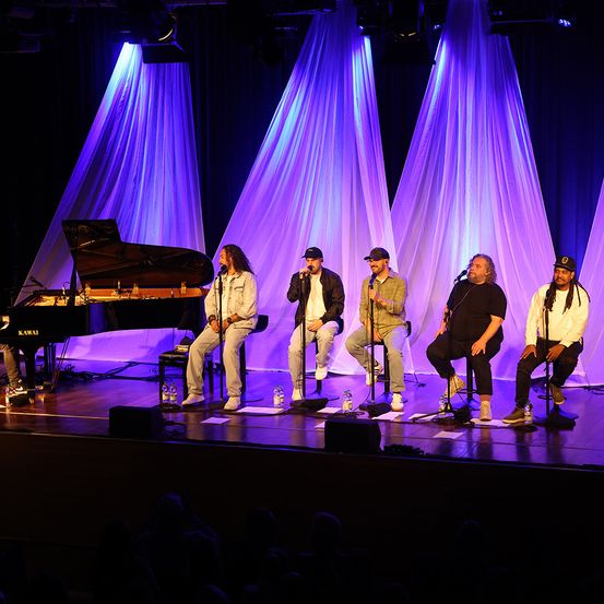 Eine Gruppe Musiker tritt auf der Bühne mit einem Flügel auf. Fünf Personen sitzen auf Hockern, jeder mit einem Mikrofon, während ein Pianist spielt. Die Bühne ist mit lila Lichtern beleuchtet und hat einen schwarzen Vorhang im Hintergrund.