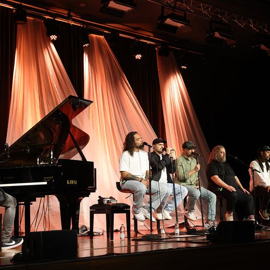 Eine Gruppe von Musikern tritt auf der Bühne auf. Ein Pianist spielt ein Konzertklavier, während mehrere Sänger in Mikrofone singen. Ein Hintergrund aus Vorhängen und Bühnenlichter vervollständigt die Szene.