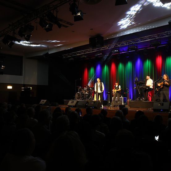 Eine Band spielt auf der Bühne vor einem farbenfrohen Hintergrund mit mehreren Scheinwerfern. Das Publikum sitzt unten.