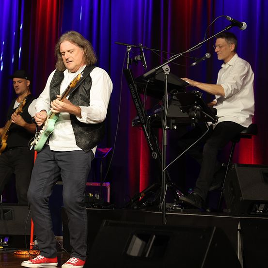 Eine Band tritt auf der Bühne auf, wobei ein Gitarrist in der Mitte steht, der eine Weste und Turnschuhe trägt. Hinter ihm spielen zwei weitere Bandmitglieder Gitarre und Keyboard. Mikrofone und Lautsprecher sind auf der Bühne positioniert.