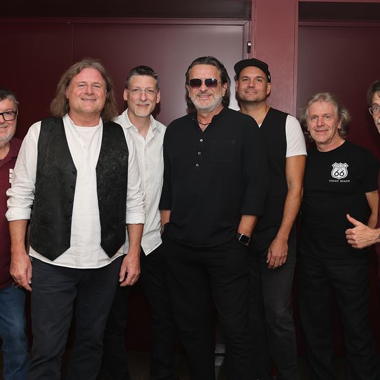 Eine Gruppe erwachsener Männer, gekleidet in Schwarz und Weiß, posiert für ein Foto in einem Raum mit einer braunen Wand.