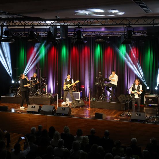 Eine Band tritt auf der Bühne auf, mit farbenfrohen Lichtern und Vorhängen im Hintergrund. Die Bandmitglieder spielen verschiedene Instrumente, darunter Gitarren, Keyboards und Schlagzeug. Das Publikum beobachtet sie aufmerksam.