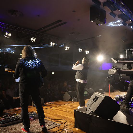 Eine Band tritt auf einer Bühne mit Scheinwerfern und Nebel auf. Der Leadsänger singt in ein Mikrofon. Ein anderer Bandmitglied spielt die Gitarre.